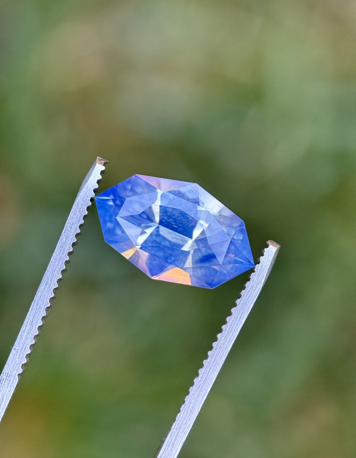 Macro view of 6.07CT periwinkle blue sapphire held in gemstone tweezers, showing opalescent light play and the detailed faceting of the precision marquise mixed cut, unheated sapphire from Sakaraha, Madagascar.