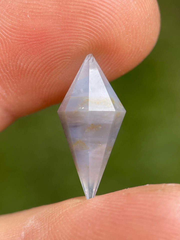 Earthy muted periwinkle sapphire weighing 8.00 carats, held between fingers, showing its elongated rhombohedral cut and unique zoning.