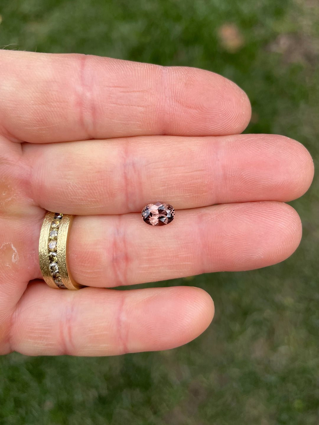 A 1.68-carat oval brilliant sapphire showing its color-shifting properties between pink and brown tones, photographed on a hand against a green grassy background.