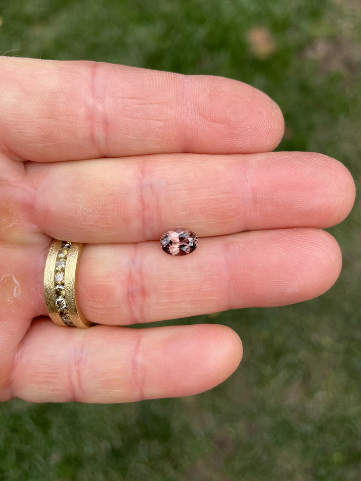 A 1.68-carat oval brilliant sapphire showing its color-shifting properties between pink and brown tones, photographed on a hand against a green grassy background.