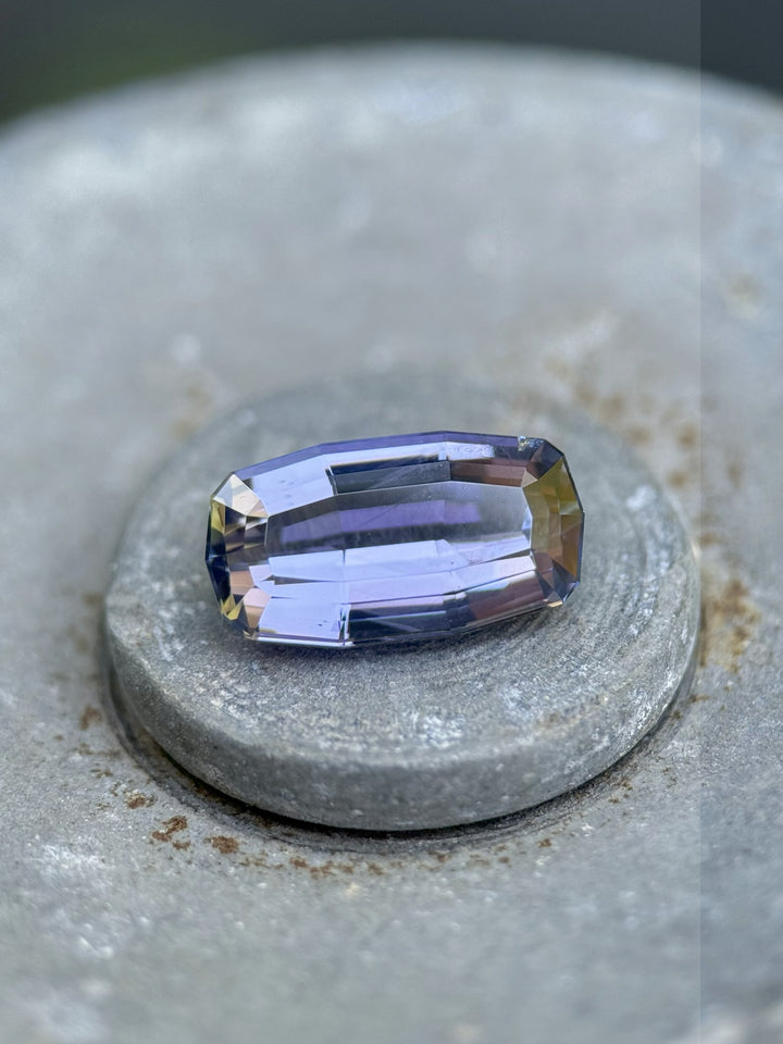 Tanzanite gemstone resting on stone surface, showing vibrant yellow, purple, and brown hues in a 2.63ct custom emerald cut