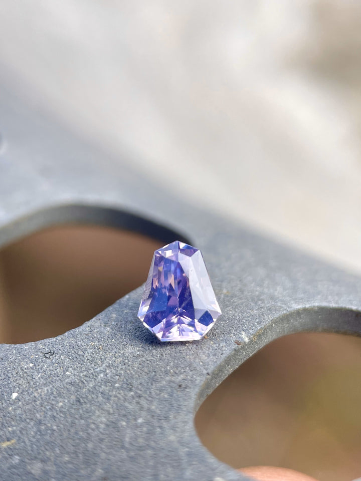Close-up of a stunning 1.14 carat sapphire with pink, lavender, and blue colors, displayed on a grey concrete surface, highlighting its unique shield cut and natural beauty.