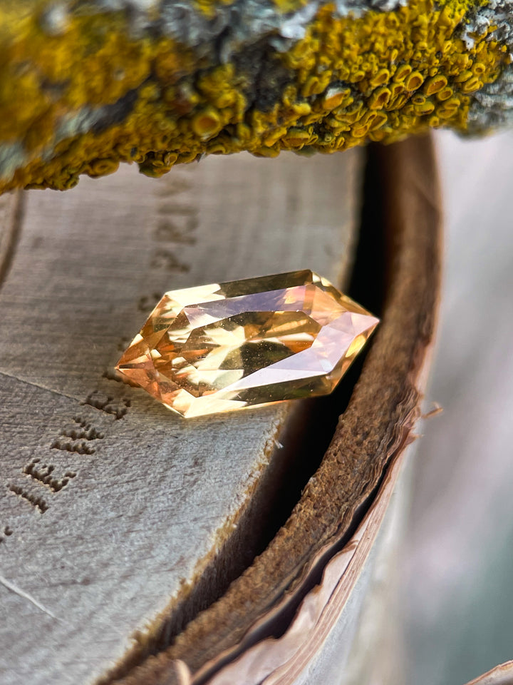 Close-up of a 2.01 carat lemon yellow sapphire in a modified brilliant hexagon cut, placed on a rustic wooden surface with moss-covered bark. Origin: Sri Lanka.