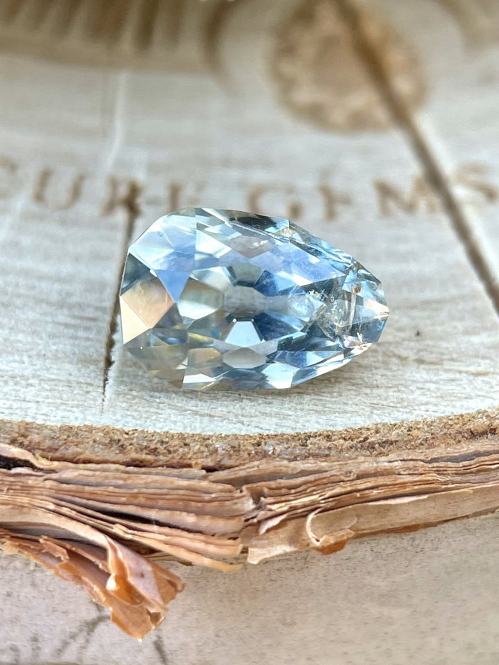 1.41-carat unheated sapphire from Malawi resting on textured wood. The pear-oval cut reveals mixed yellow, green, and blue tones under natural light, showcasing its pastel parti color palette.