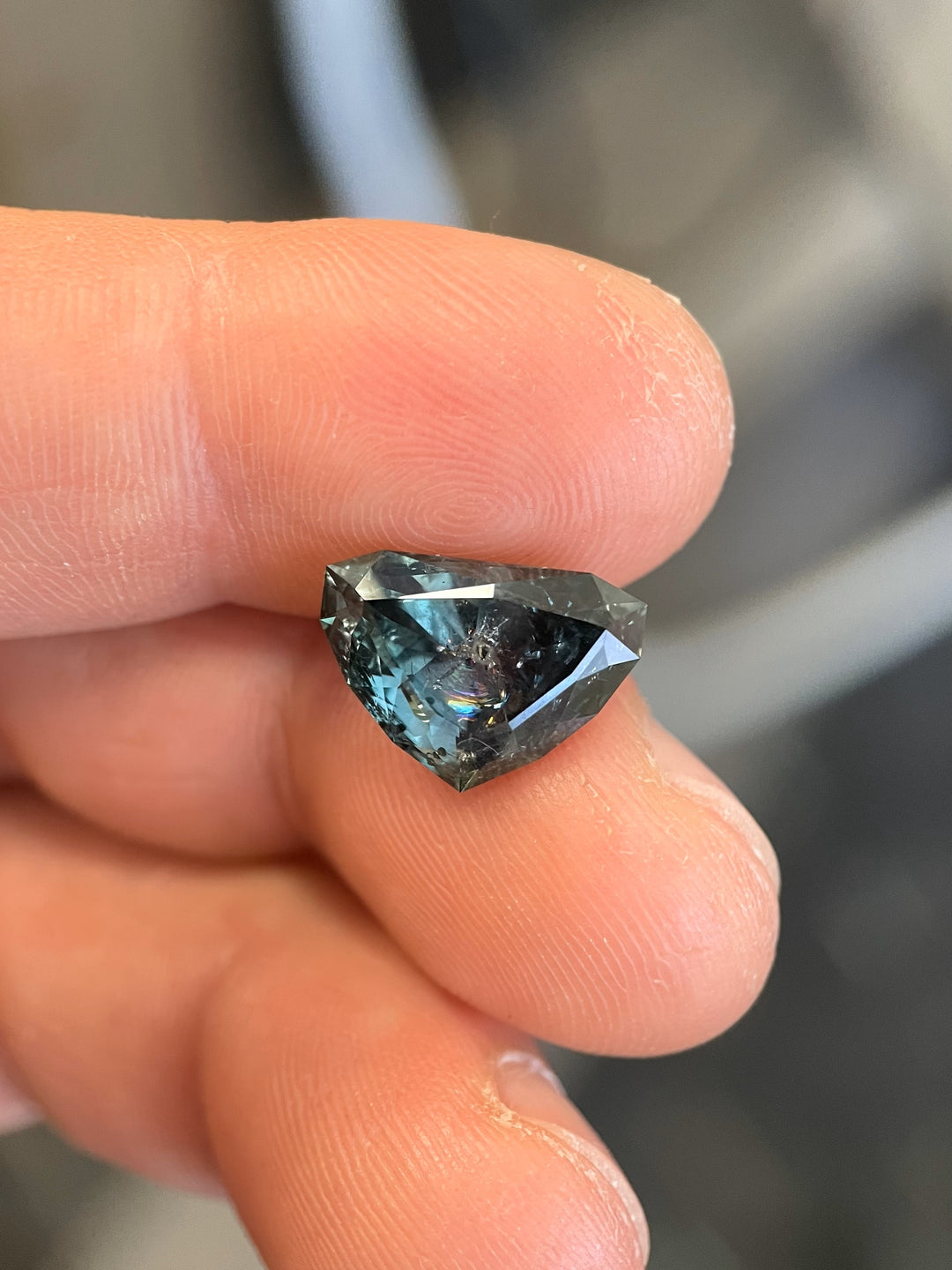 A 7.06-carat natural sapphire showcasing a bluish-green hue with a color shift to gray-purple, held against a green background. Modified shield brilliant cut.