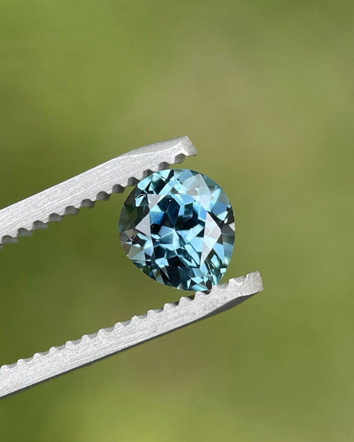 Close-up of 1.02ct modified brilliant pear cut Montana sapphire, teal-blue color, held in gem tweezers.