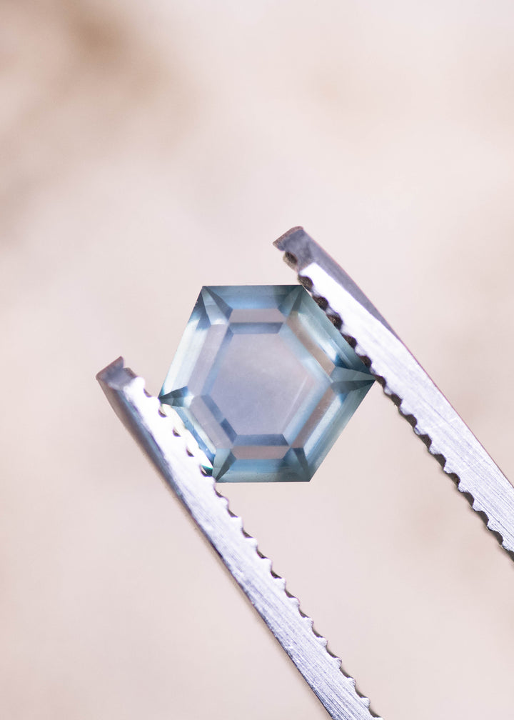 Geometric hexagonal step cut teal blue seafoam sapphire held in gem tweezers, highlighting the precision cut facets, light performance, and untreated Montana sapphire clarity at 1.37 carats.