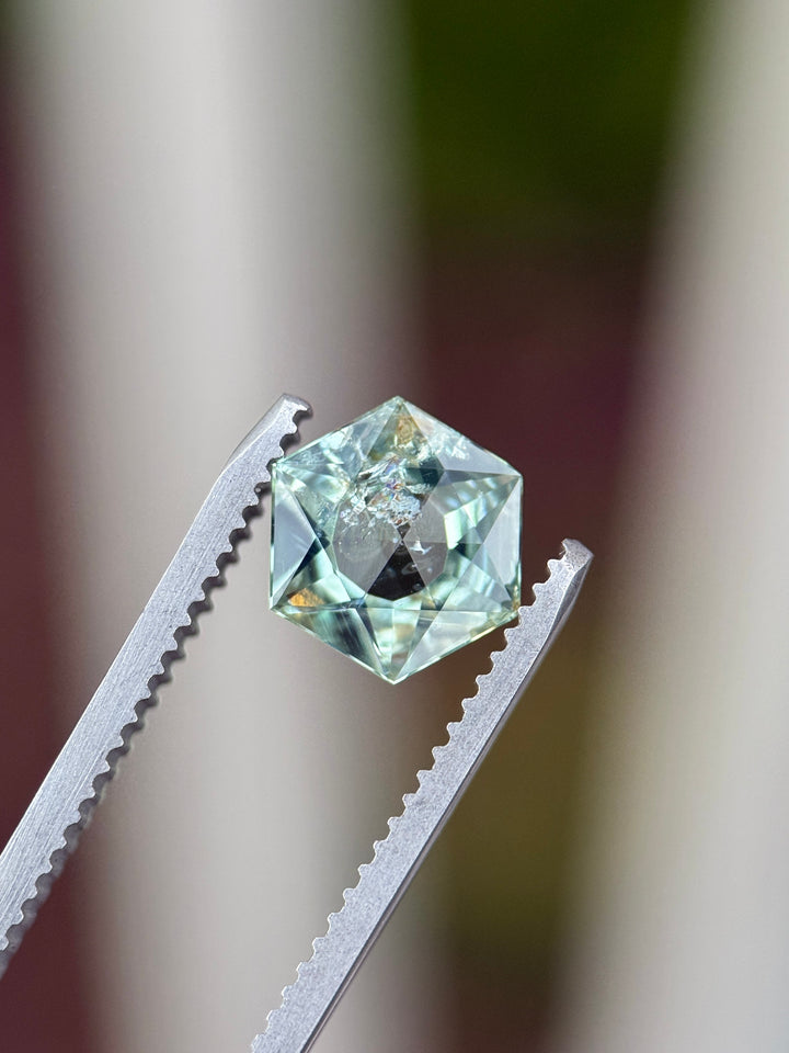 Precision cut Montana sapphire held in jeweler’s tweezers, displaying a custom hexagon mixed cut with rose cut table, open culet, and mint sage teal green color in a 1.76 carat untreated sapphire
