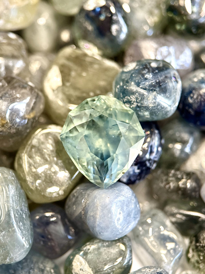 Natural unheated Montana sapphire, 7.08 carats, in a custom pear shape, placed among raw sapphire pebbles.