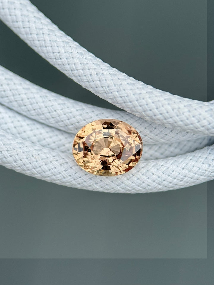 Natural 1.51ct oval sapphire in soft peach and pink hues, displayed on white braided cord for texture contrast