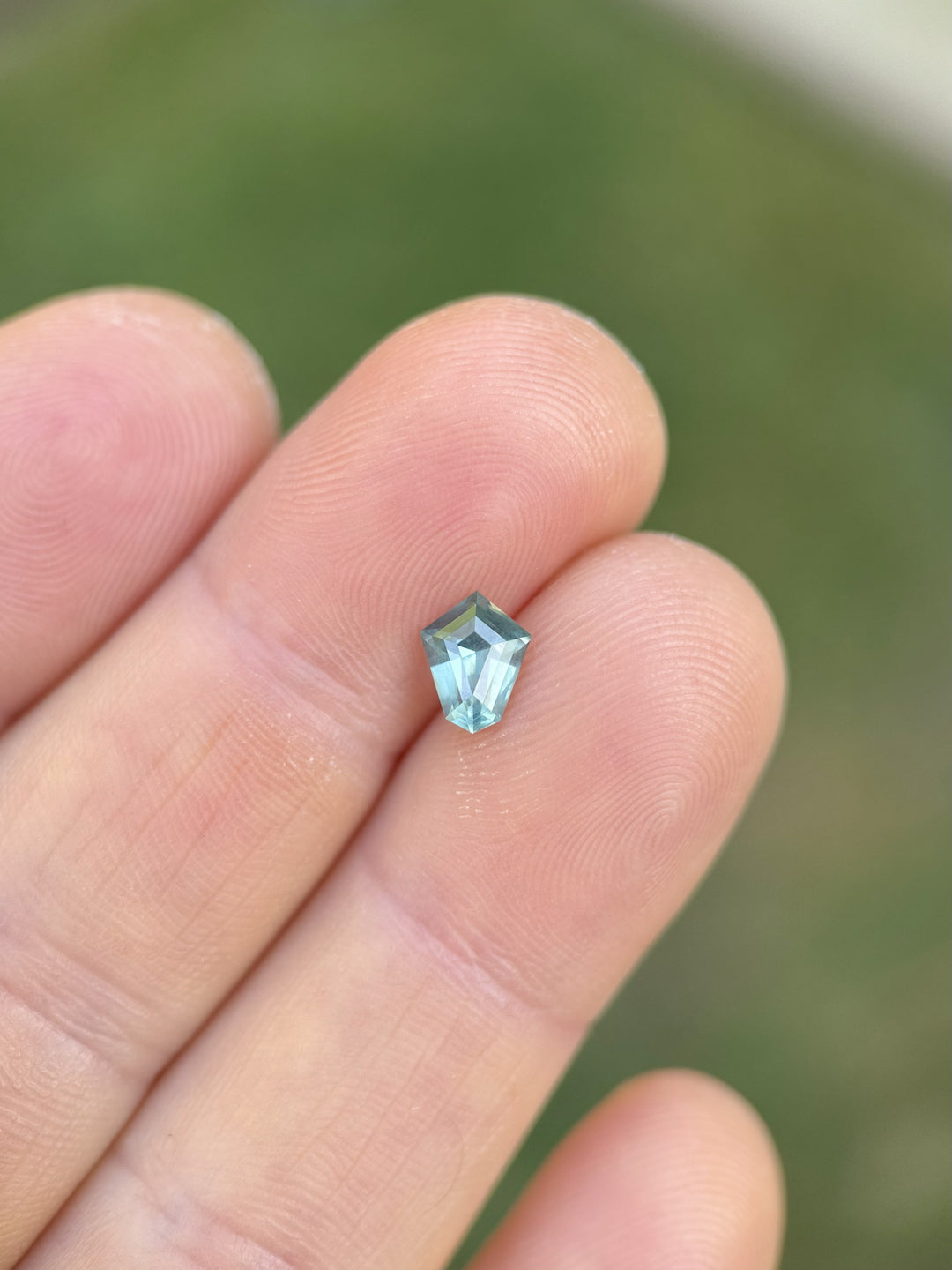 Teal and green parti sapphire from Montana, 1.14 carats in a custom step cut shield shape, displayed on fingertips against a blurred grassy background for scale and natural color contrast.