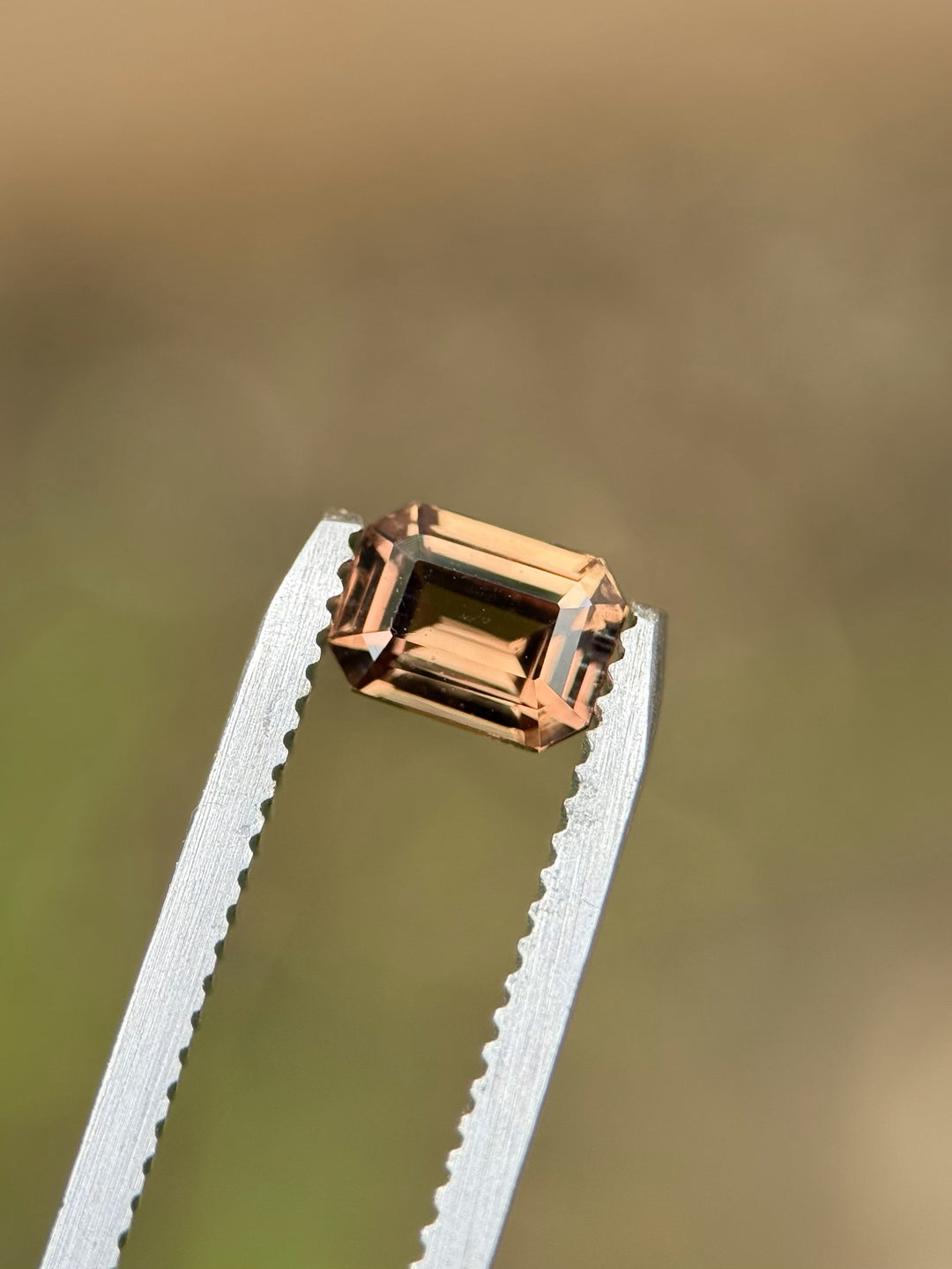Precision-cut 1.10ct sapphire in peach and orange tones held by gem tweezers against a soft neutral background.