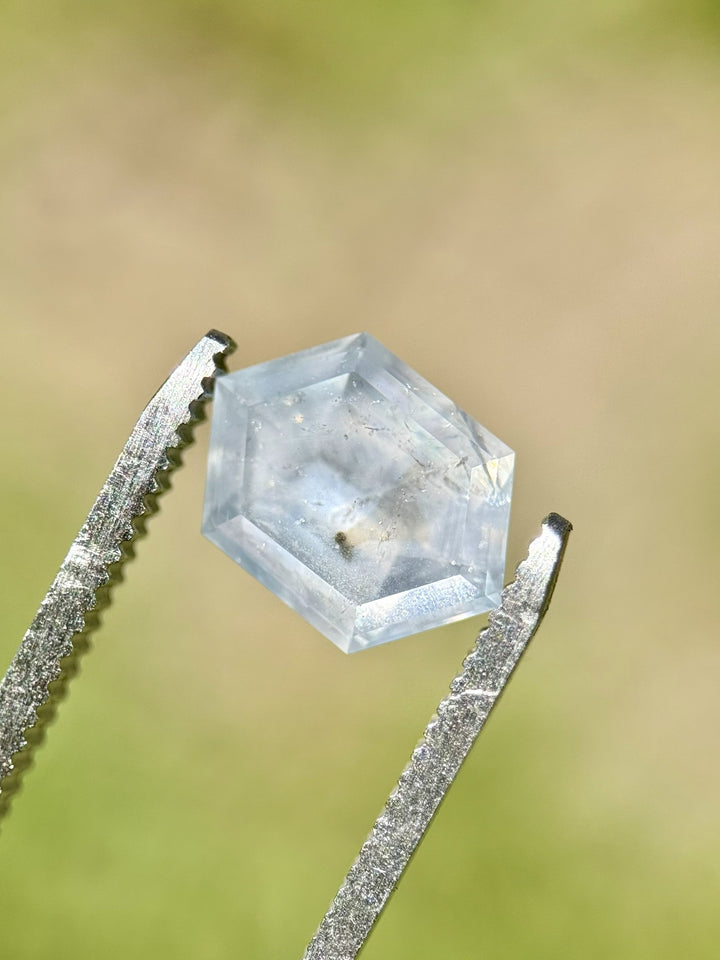 Natural grayish lavender Montana sapphire held in gem tweezers outdoors, showing hexagon step cut and surface clarity in direct light