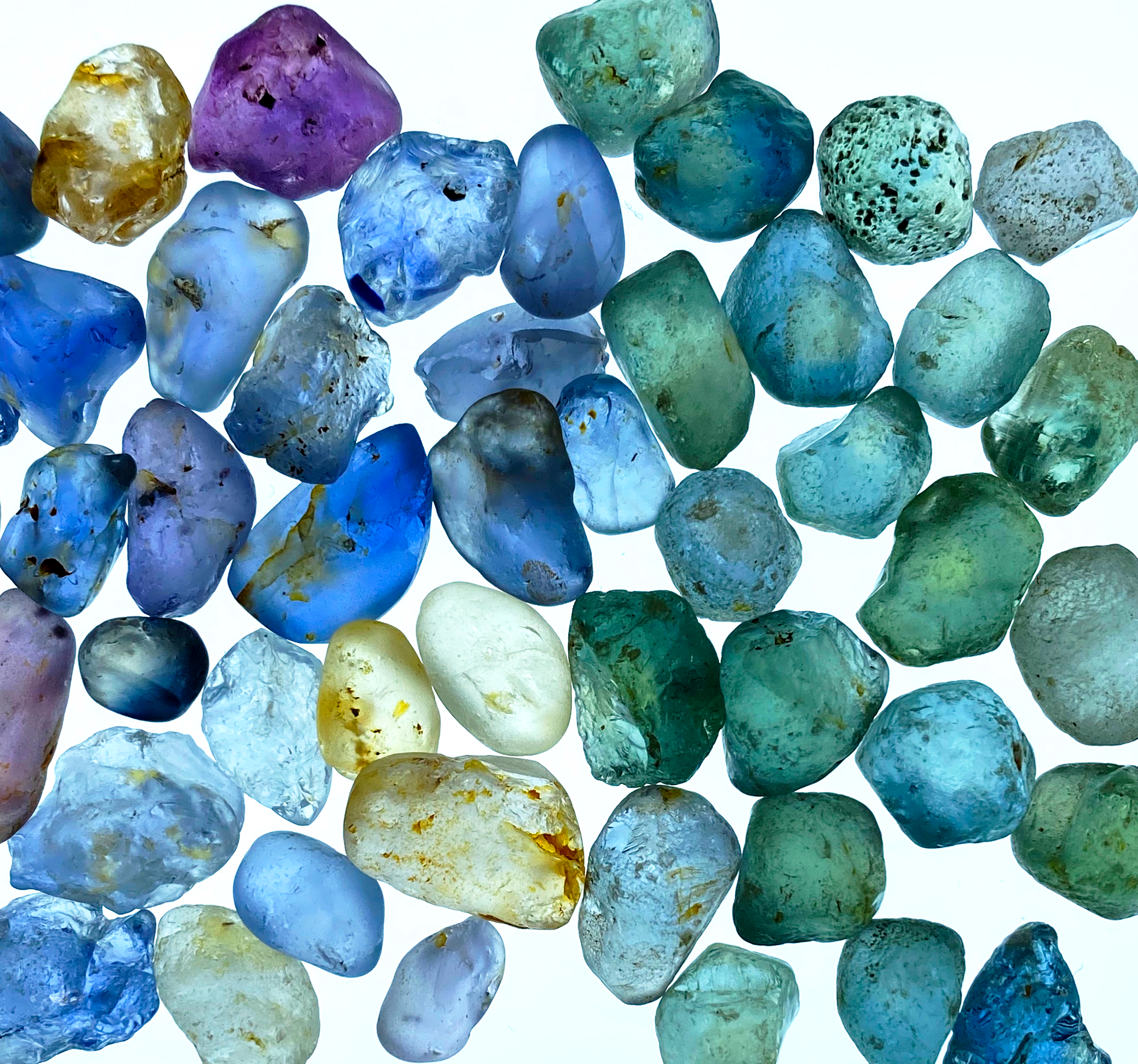 Natural rough sapphires in blue, green, teal, purple, and yellow tones, showcasing raw gemstone color before cutting.