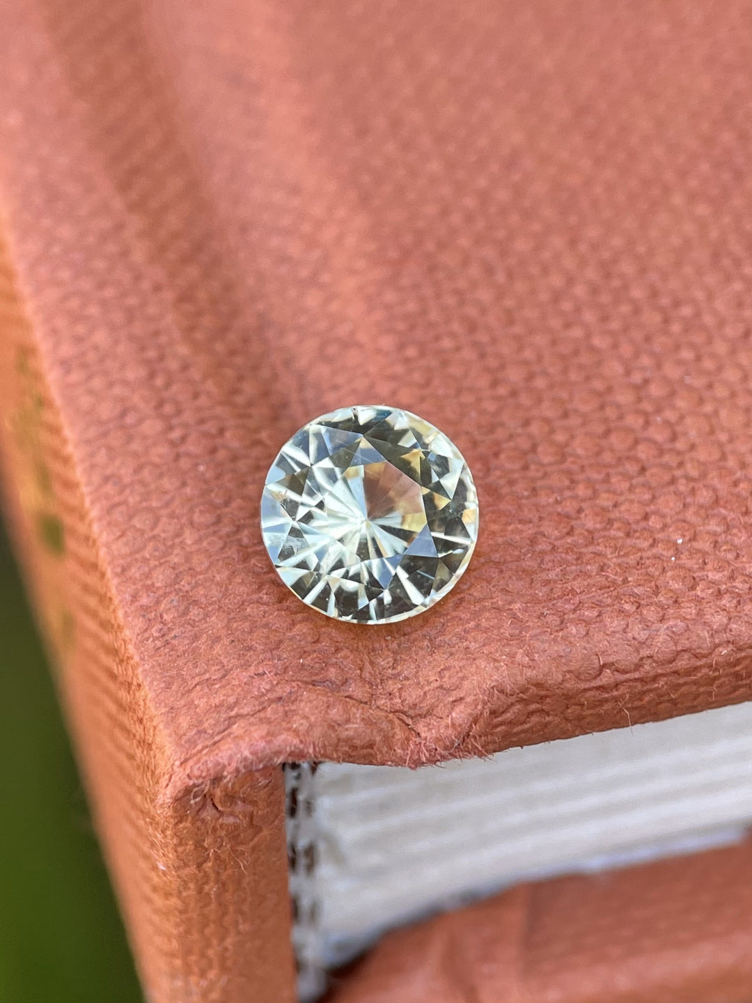 A 1.59-carat peach yellow sapphire with a round brilliant cut, displayed on a textured book surface, showcasing its vibrant sparkle and clarity.