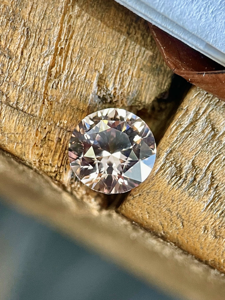 A 2.57-carat round brilliant peach sapphire placed on rustic wood, capturing its soft color and symmetrical faceting in natural light.