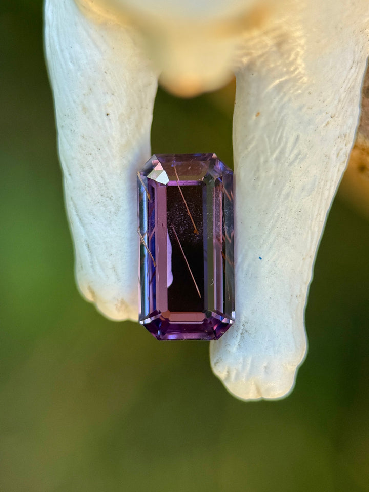 Rutilated purple sapphire displayed between organic bone elements, showcasing elongated emerald portrait cut, sharp step facets, internal rutile needles, and natural untreated color from Madagascar