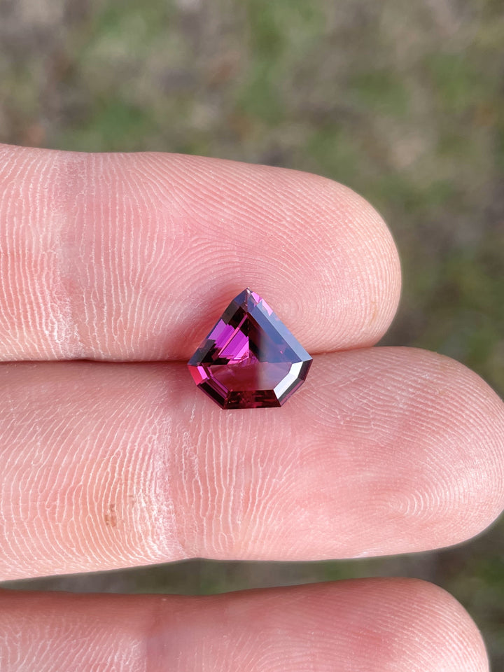 A striking shield step cut garnet in purple and pink tones, weighing 2.61 carats, resting on a mineral background.