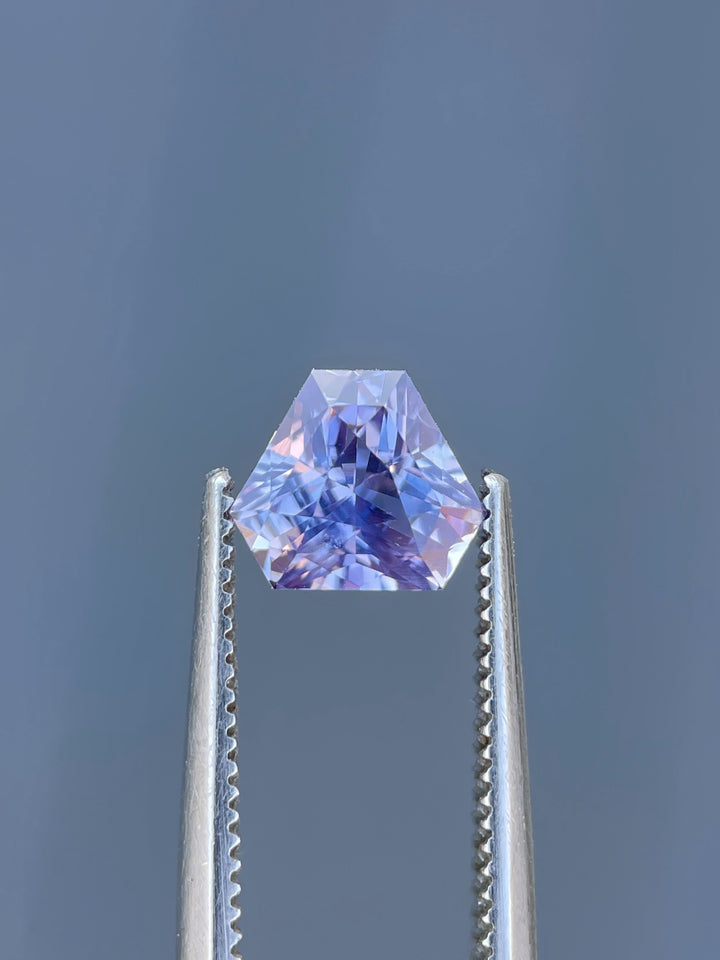 A stunning lavender sapphire, 1.56 carats, held in gemstone tweezers against a gray background, highlighting its intricate triangle cut and vibrant sparkle.