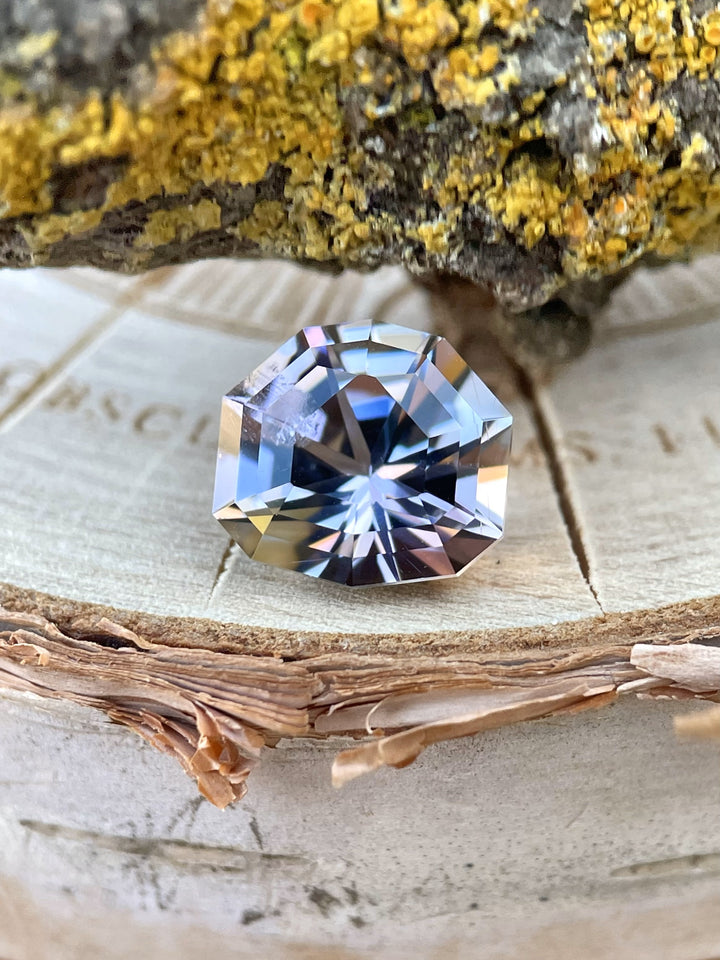3.33-carat Tanzanite with custom step cut shield resting on natural wood and lichen, displaying pastel mermaid, gray, and lavender tones from Merelani Hills, Tanzania
