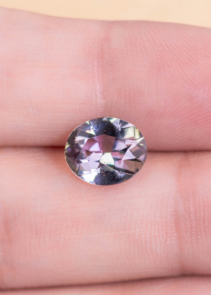 Natural 2.57 carat Tanzanite resting on a finger for scale, displaying a custom modified brilliant oval cut with subdued teal blue and lavender tones, untreated and sourced from Merelani Hills, Tanzania