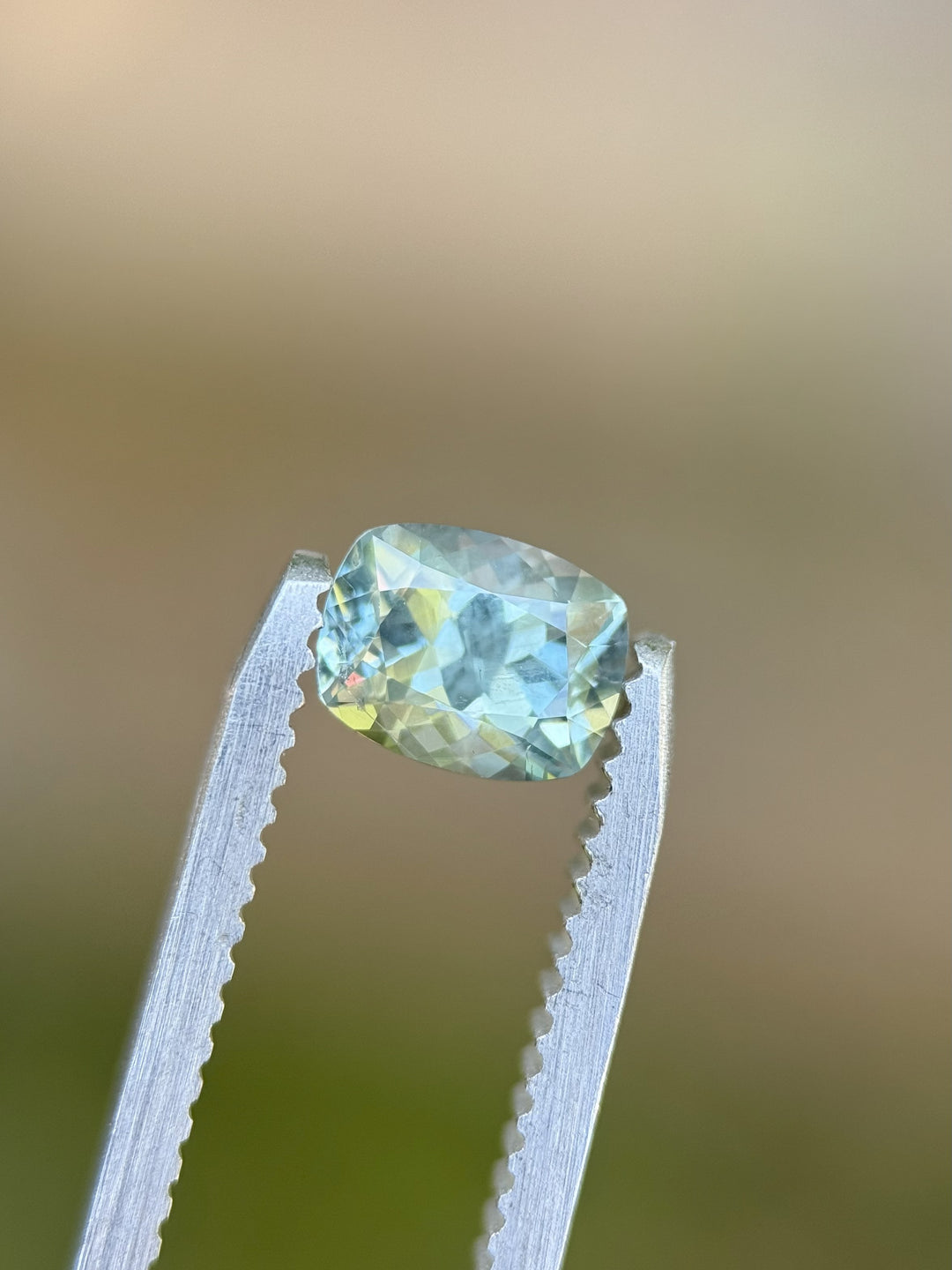 Natural Montana sapphire in elongated cushion cut held in tweezers, displaying teal, green, and blue hues, 1.15 carats unheated