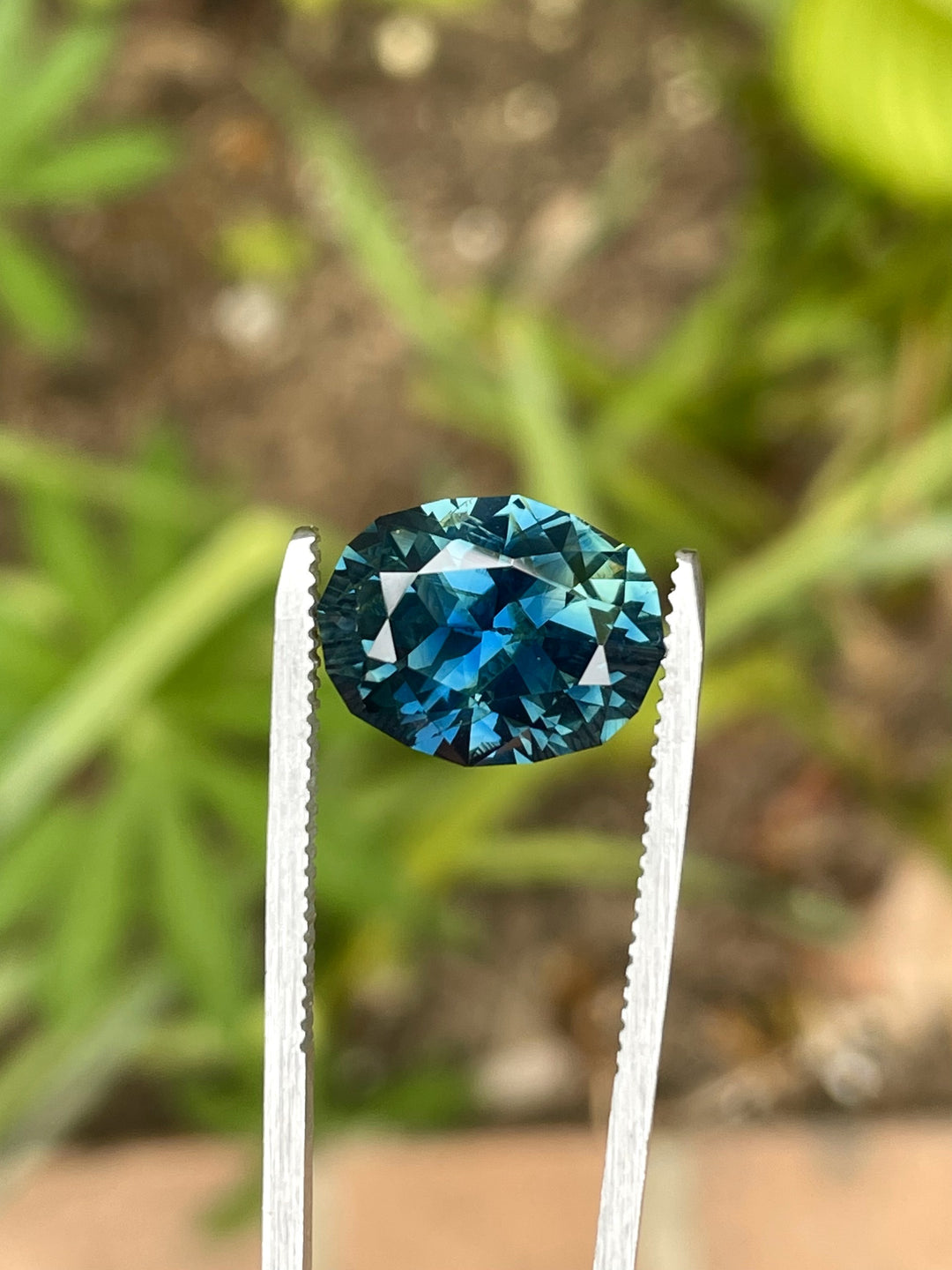 Natural untreated teal-blue sapphire weighing 7.75 carats, held in gemstone tweezers with blurred green garden background.