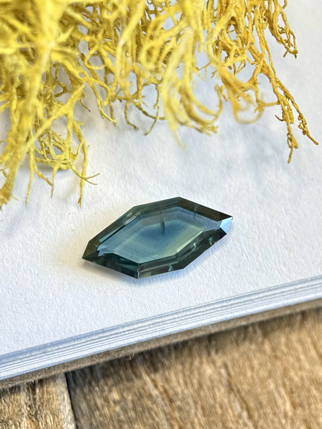 Freeform sapphire placed on white paper with soft light highlighting its deep blue and teal saturation.