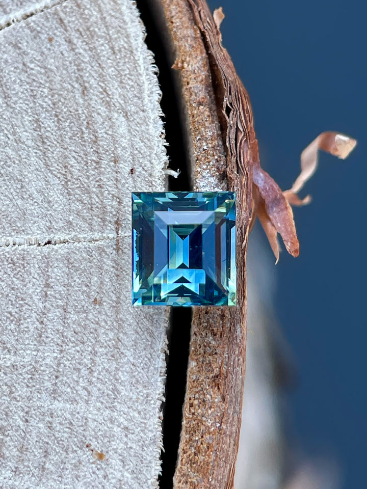 A mesmerizing bi-color sapphire in green and blue, featuring a precise antique-style Carre cut, resting on a wooden surface.
