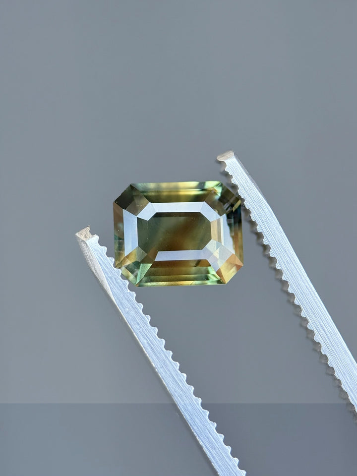 2.02ct untreated sapphire in emerald cut held by gem tweezers, showing color zones of orange, green, and grey from Garba Tula, Kenya.