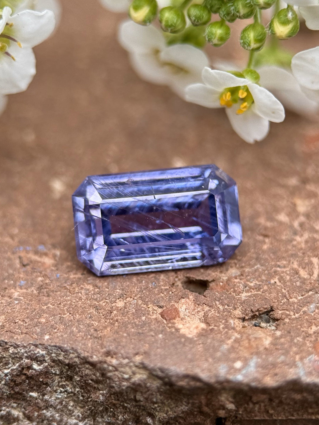 Natural violet Umba sapphire resting on rustic surface with white flowers, showing brilliance and antique-style emerald shape