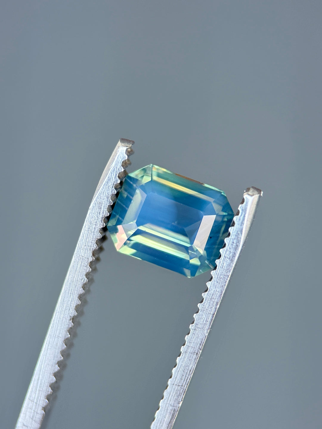 Natural unheated opalescent sapphire held in tweezers, highlighting yellow, teal, blue, and green color transitions across the gemstone.