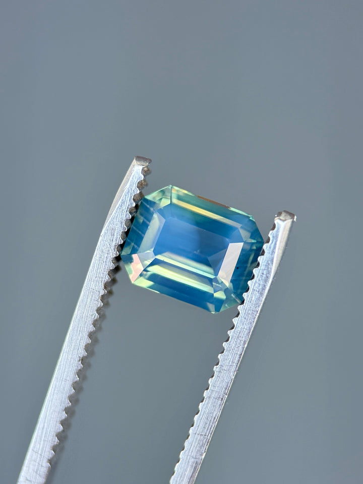Natural unheated opalescent sapphire held in tweezers, highlighting yellow, teal, blue, and green color transitions across the gemstone.