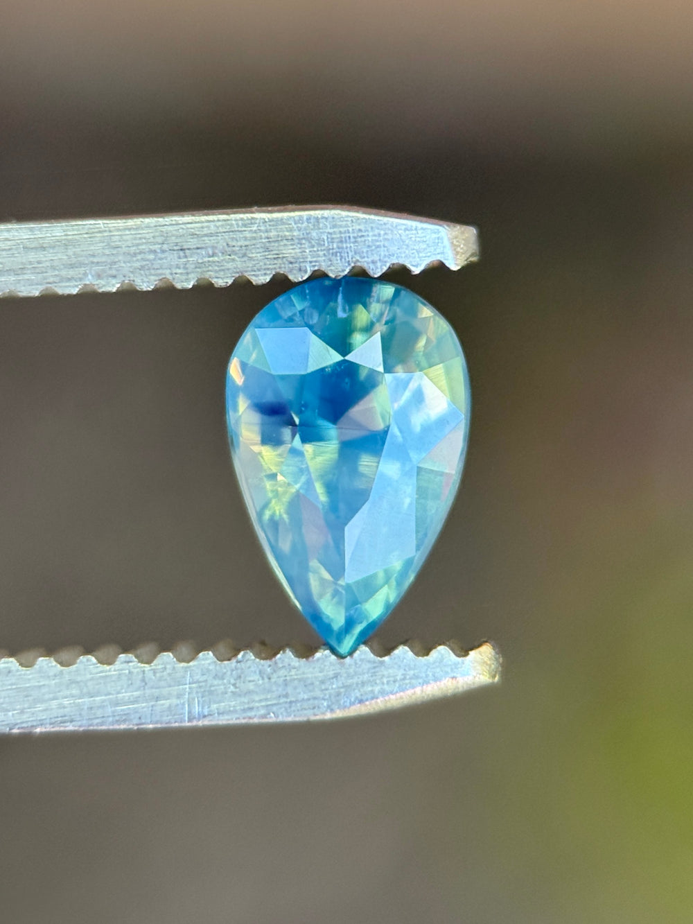 Natural unheated teal blue green sapphire held in tweezers, highlighting the elegant pear shape and subtle opalescent glow.