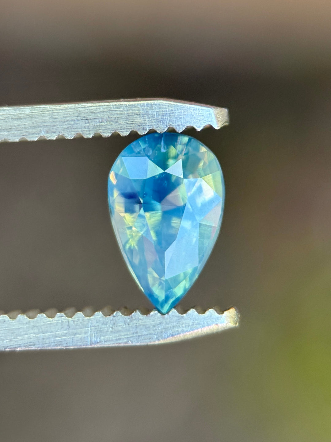 Natural unheated teal blue green sapphire held in tweezers, highlighting the elegant pear shape and subtle opalescent glow.