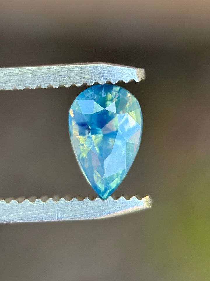 Natural unheated teal blue green sapphire held in tweezers, highlighting the elegant pear shape and subtle opalescent glow.