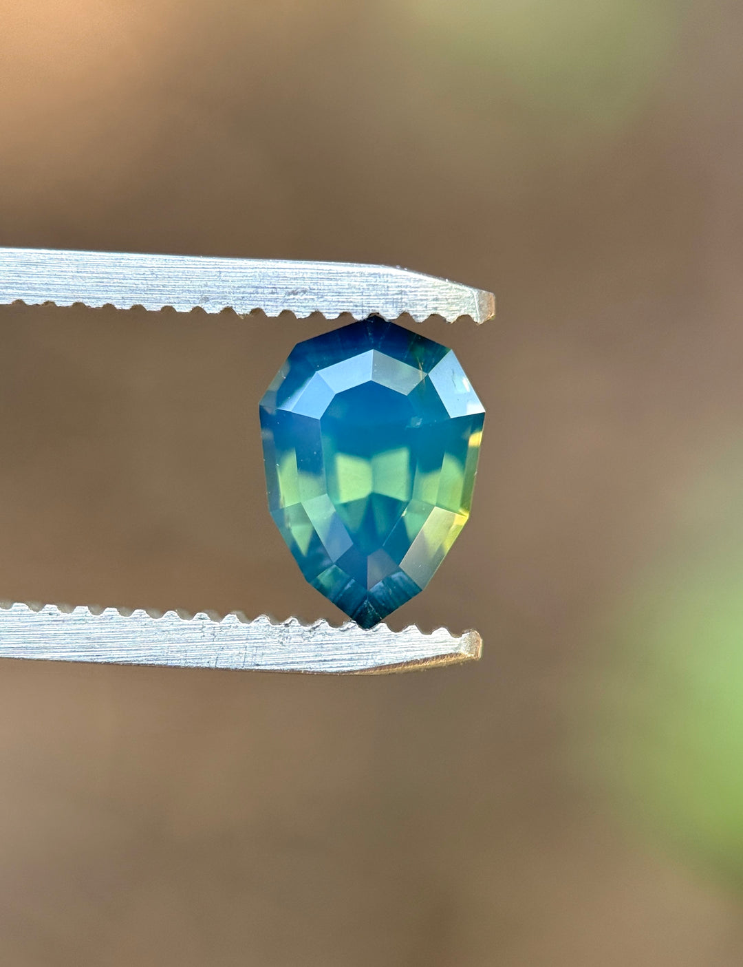 Natural unheated teal blue green sapphire held in tweezers, highlighting the geometric shield outline and reflective step-cut facets.