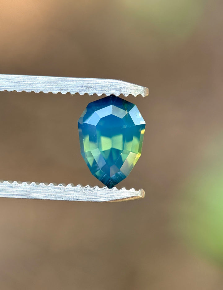 Natural unheated teal blue green sapphire held in tweezers, highlighting the geometric shield outline and reflective step-cut facets.