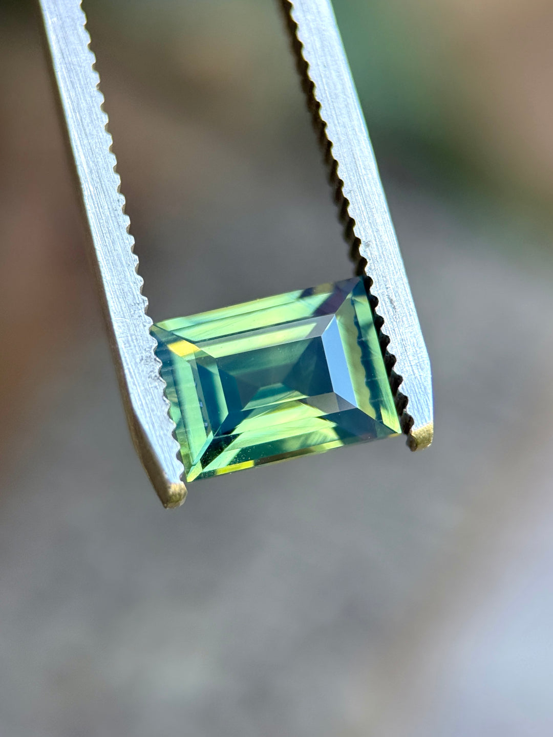 Natural unheated teal blue green sapphire held in tweezers, highlighting crisp step-cut geometry and opalescent glow.