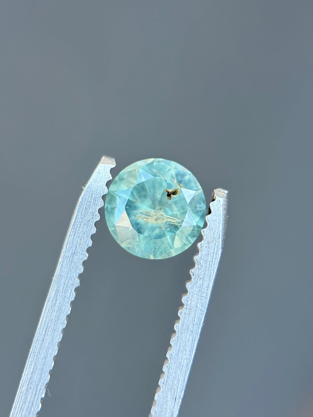 Natural untreated powder teal sapphire held in tweezers, highlighting round brilliant faceting and luminous pastel teal color.