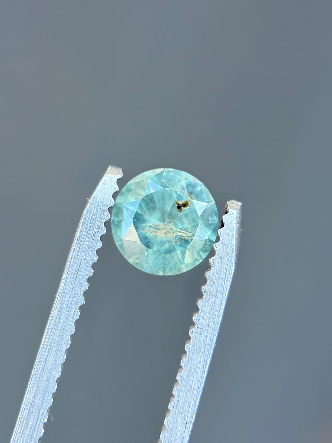 Natural untreated powder teal sapphire held in tweezers, highlighting round brilliant faceting and luminous pastel teal color.