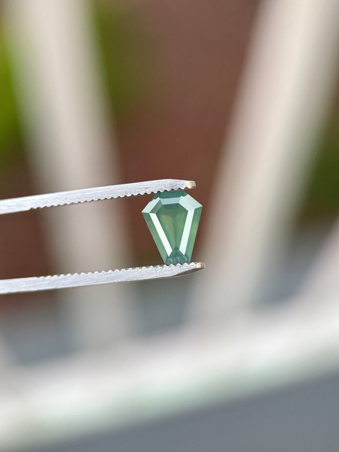 Natural untreated teal blue green sapphire held in tweezers, highlighting the geometric shield shape and reflective step-cut facets.