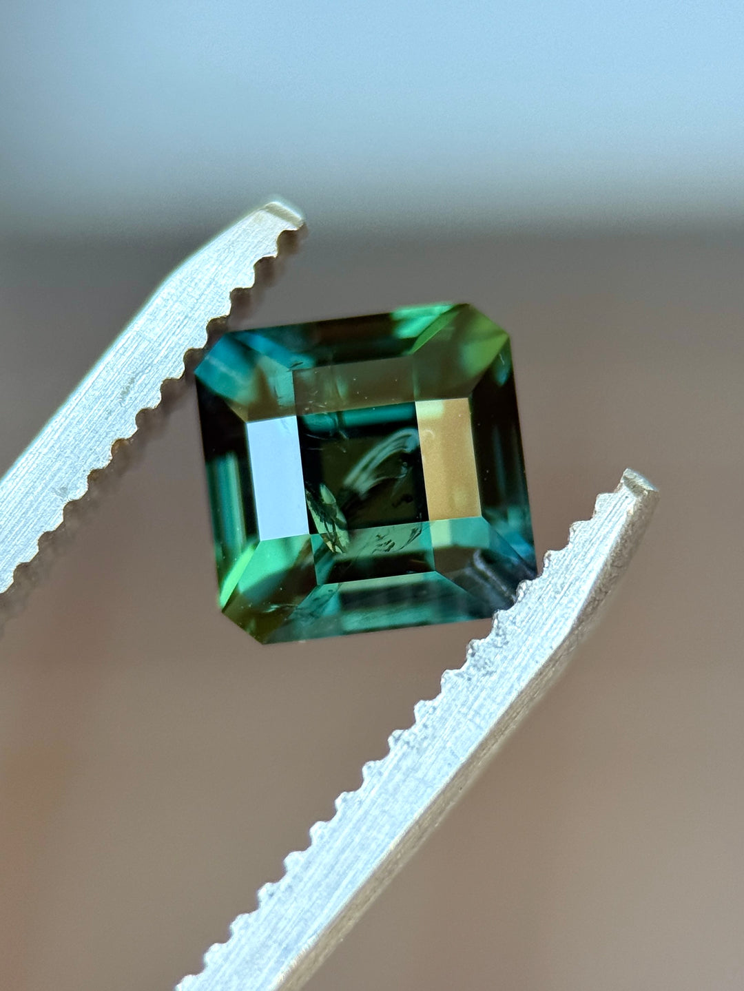 Natural untreated teal green sapphire held in tweezers, highlighting the geometric antique emerald cut and hall-of-mirrors reflections.