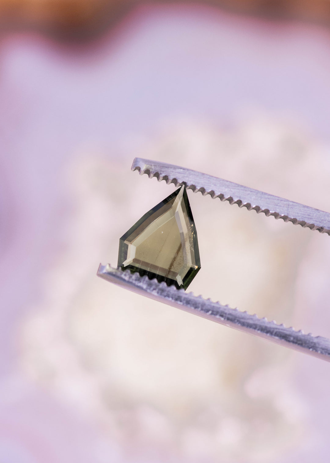 Loose olive green sapphire held in tweezers, displaying the custom shield portrait cut and step-style facet structure from Nigeria.