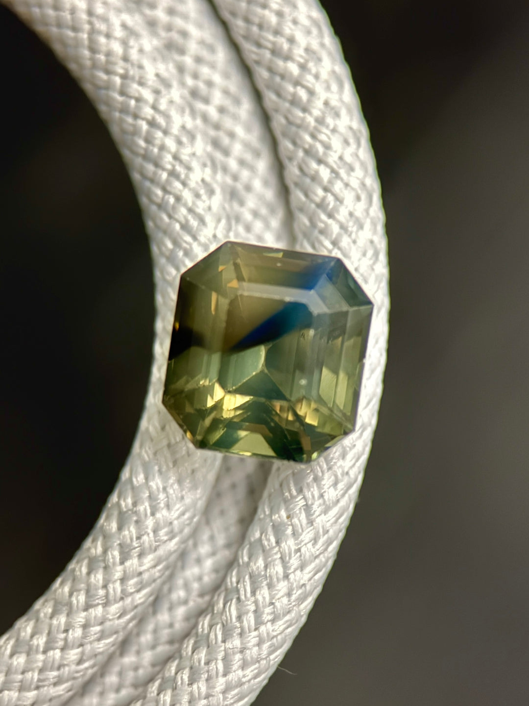 3.40 carat opalescent sapphire resting on white rope, precision emerald cut sapphire showing earthy olive, teal, green, brown, and blue tones from Gombe, Nigeria