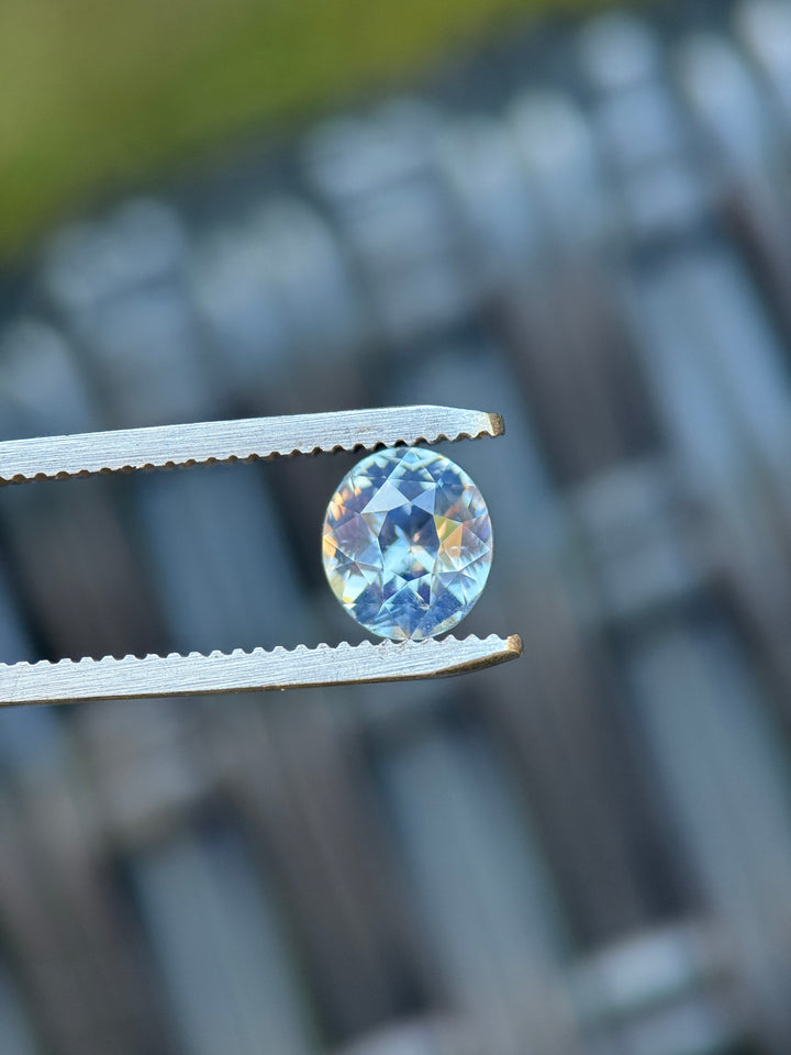 2.53ct opalescent cornflower blue oval sapphire held in silver gem tweezers, showing crisp triangular and kite-shaped crown facets with soft glowing blue body color and pastel flashes.