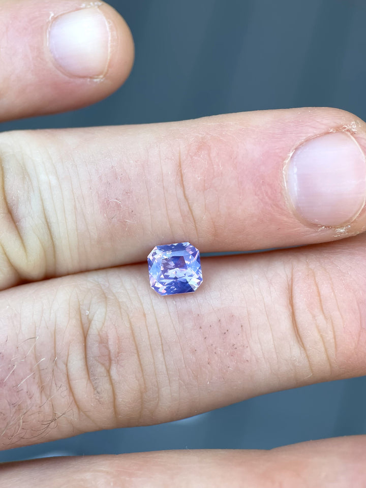1.57 carat sapphire held between fingers, emphasizing its custom radiant cut and stunning pastel shades of pink and purple.