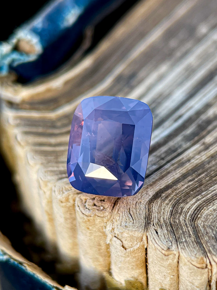 A 2.56-carat cushion-cut sapphire with mauve, pink, and purple hues, displayed against a rustic background.