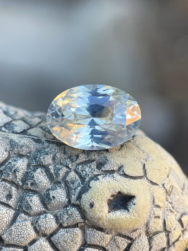 Close-up view of a 4.23-carat opalescent white sapphire in an oval brilliant cut, showcasing its white and yellow hues on a textured surface.