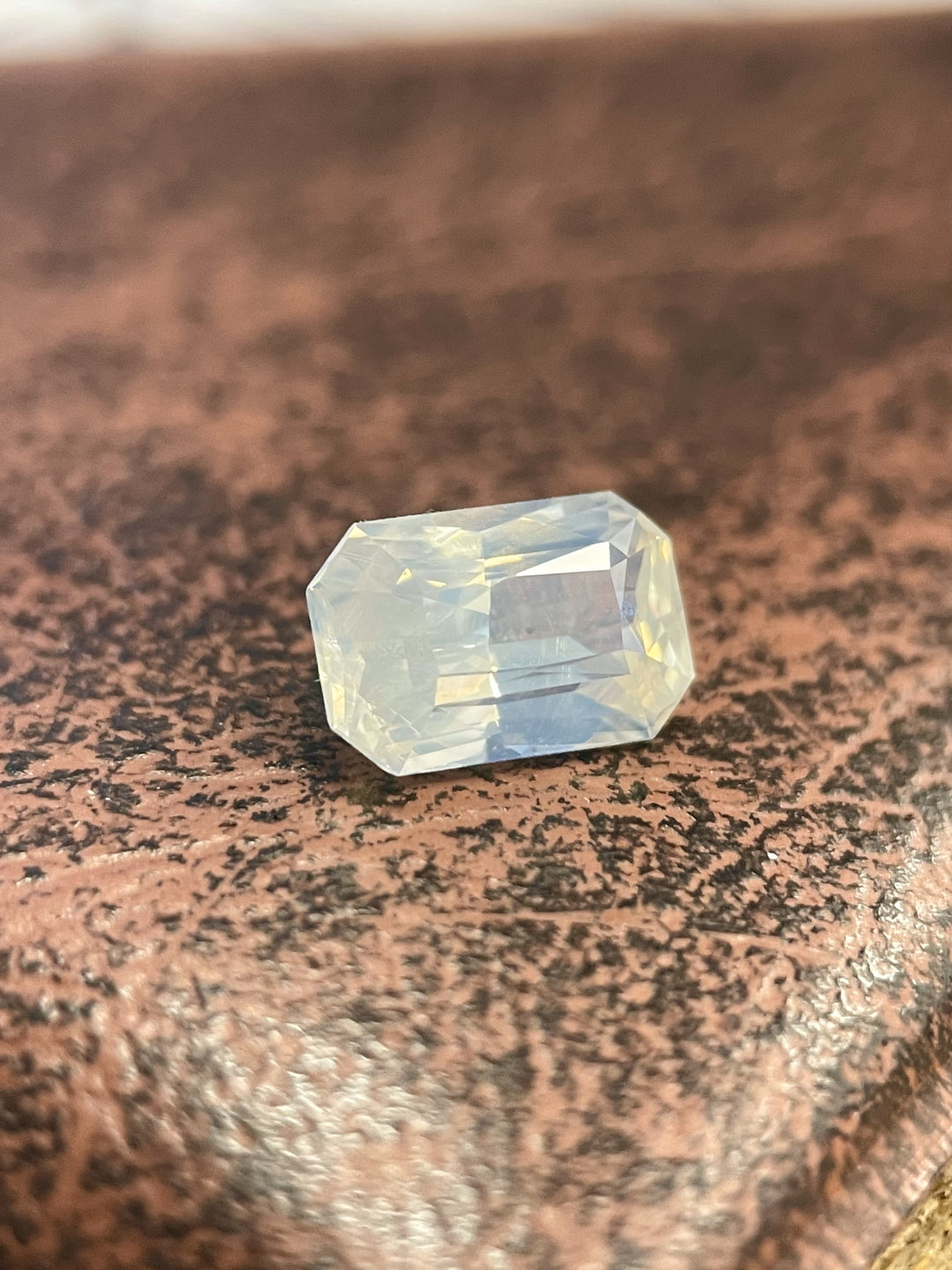 An opalescent yellow and gray sapphire weighing 2.10 carats, cut in an elongated radiant shape. The gemstone is set against a textured brown surface, showcasing its shimmering hues and precise faceting. Dimensions: 8.80mm length, 5.80mm width, 4.30mm height